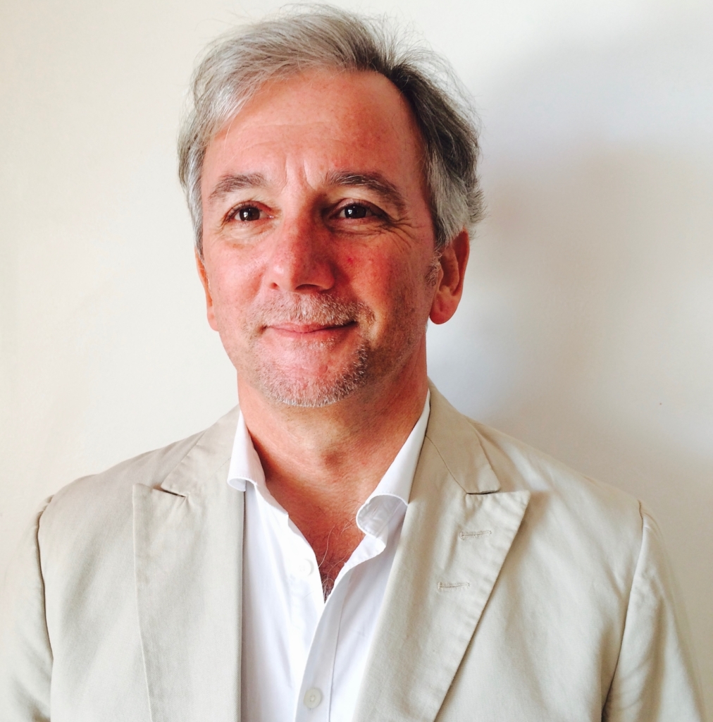 A portrait photograph of the author, Michael Emmerich, smiling whilst wearing a cream coloured suit with a white shit.
